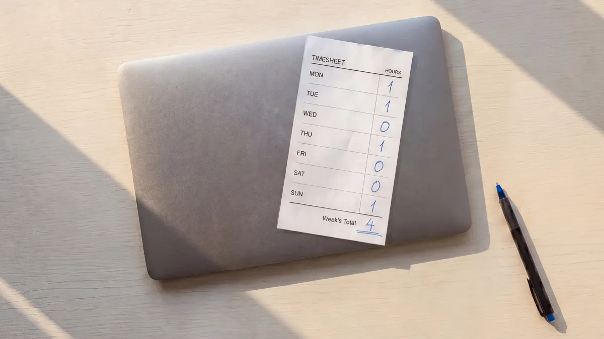 A printed timesheet showing a four-hour weekly total lies partly on a closed laptop, beside a pen on a sunlit desk.